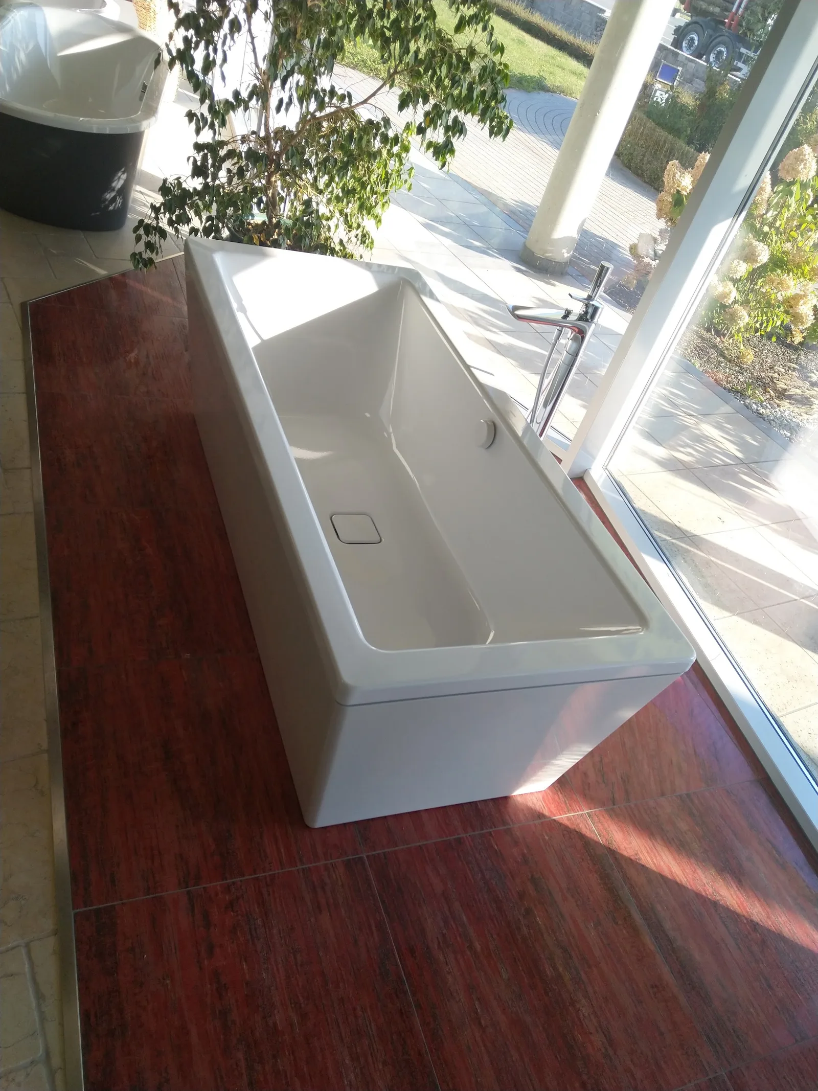 Freistehende weiße Badewanne auf dunkelrotem Boden vor einer Glasfront im Schauraum
