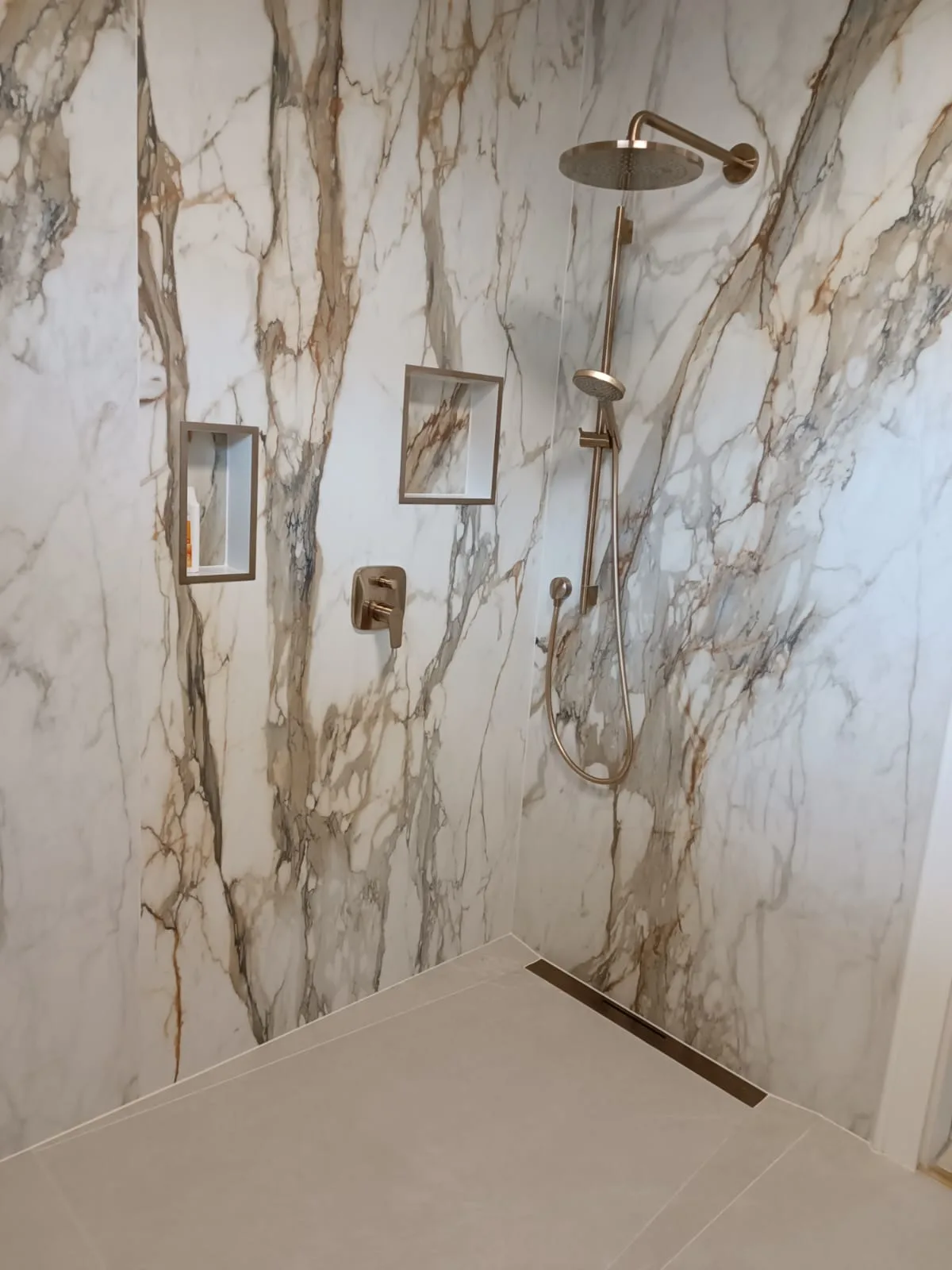 Bodengleiche Dusche mit großformatigen Wandplatten in heller Marmoroptik und goldfarbener Armatur