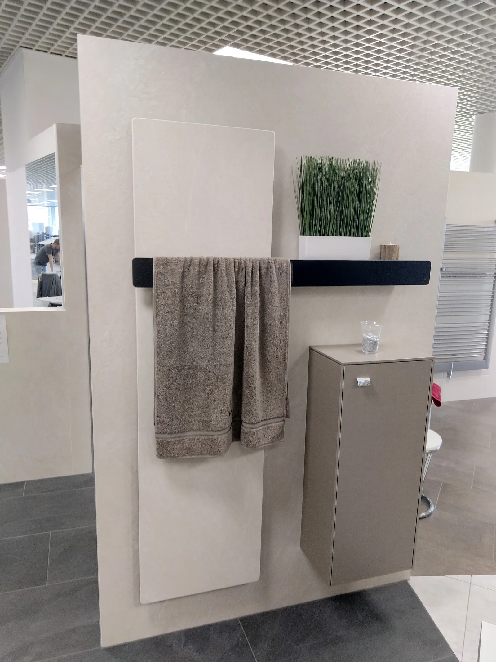 Helle Wandplatte mit schwarzer Handtuchstange, grauem Handtuch und kleinem Badschrank im Schauraum